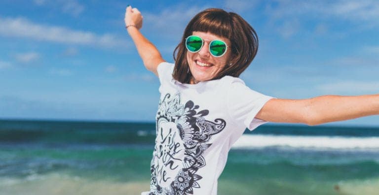 Brunette woman wearing green reflective sunglasses and a white t-shirt jumps happily on a beach