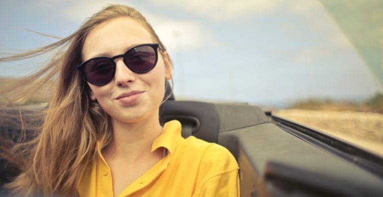 Headshot of a smiling blonde woman wearing sunglasses and a yellow blouse while sitting in the back seat of a convertible