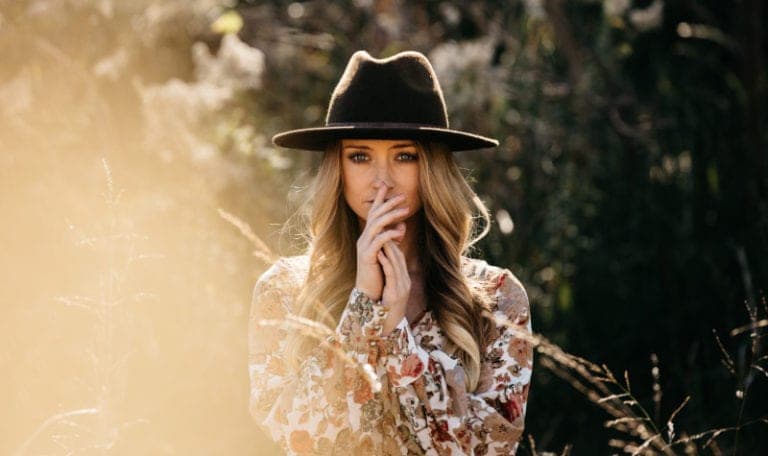 Blonde woman wearing a floral blouse and a dark hat covers her mouth with her fingers due to recurring halitosis