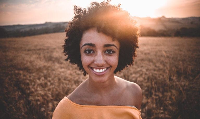 Curly-headed woman wearing a yellow blouse smiles with strong enamel while standing in a wheat field at sunset
