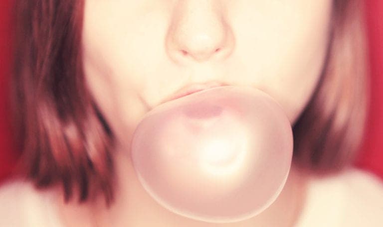 Closeup of a woman with brown hair blowing a pink gum bubble against a red wall
