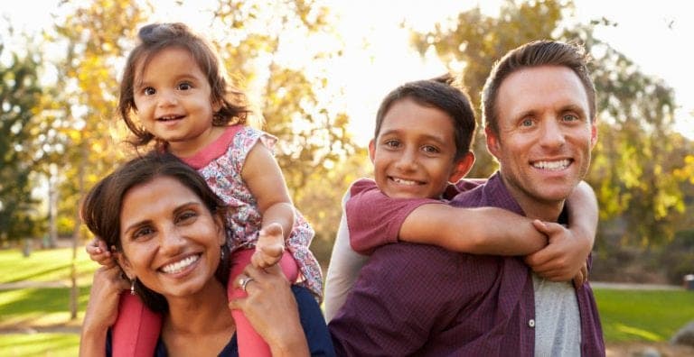 A husband and wife smiling in the sunlight as they play with their kids on their backs