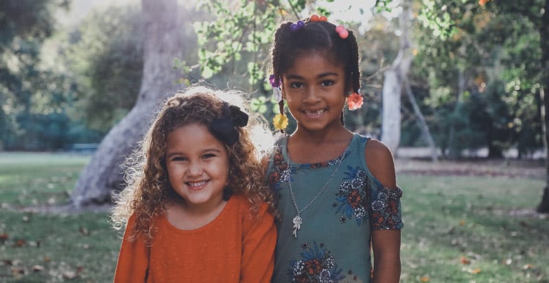 A young girl with curly hair stands outside next to a dark-haired with braids missing her two front teeth