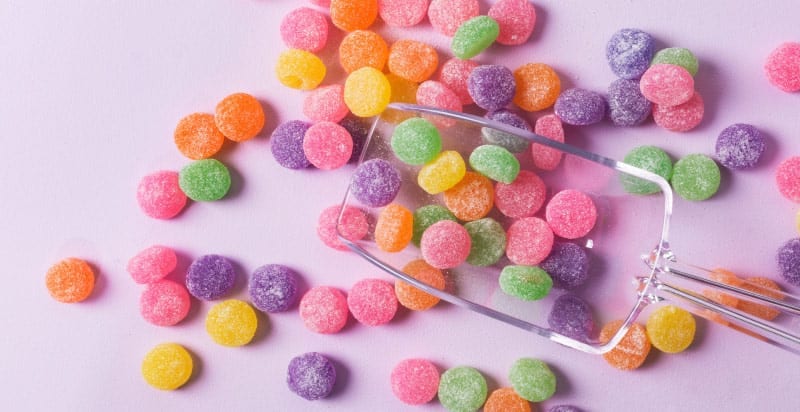 Aerial view of yellow, pink, orange, green, and purple sugary and sticky gumdrops on a lavender counter with a clear scooper
